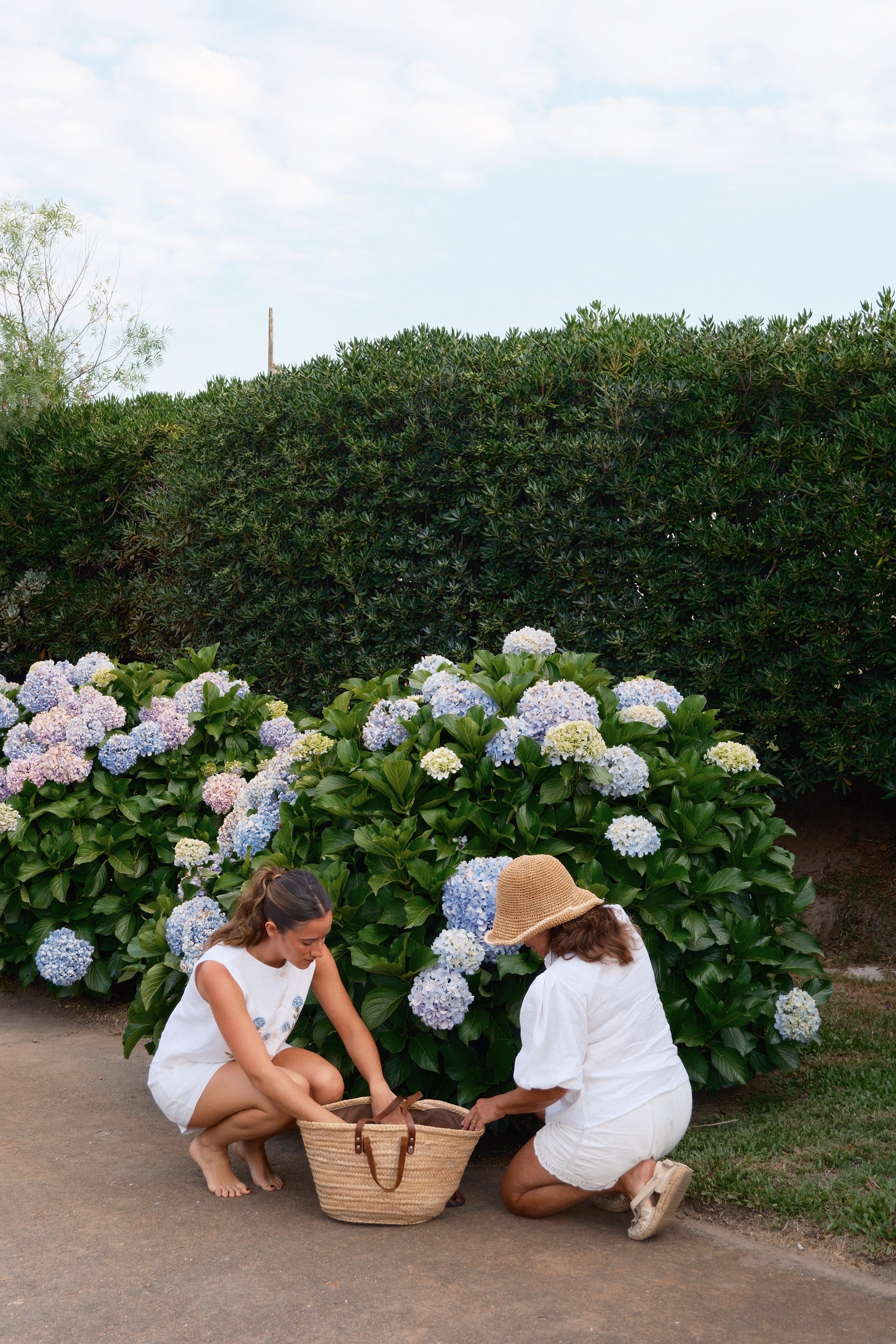 Two women in white dresses and hats interacting with a large woven basket in front of hydrangea bushes.
